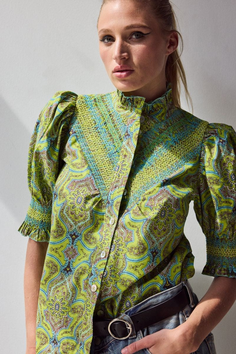 A woman with blonde hair in a ponytail wears the HOPE FOR FLOWERS Short Sleeve Smocked Shirt in Green Foulard, made of organic cotton, paired with faded blue jeans, standing with her back to the camera against a plain light background.