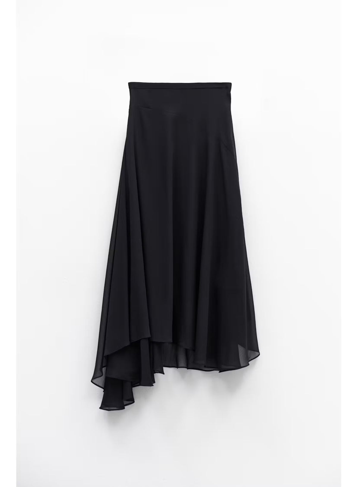 A woman stands indoors by large windows, showcasing a full-length silhouette in the ELEH Asymmetrical Skirt in Black, paired with a black short-sleeve top and black ankle boots.