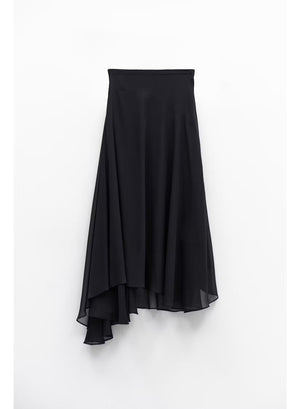 A woman stands indoors by large windows, showcasing a full-length silhouette in the ELEH Asymmetrical Skirt in Black, paired with a black short-sleeve top and black ankle boots.