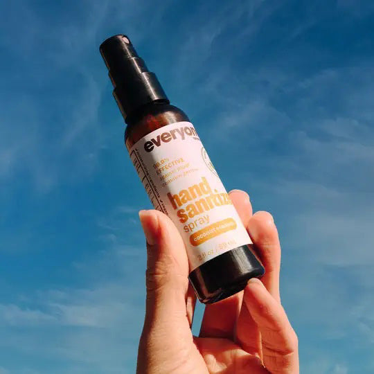 A brown leather bag is open, showing a bottle of Everyone For Everybody Coconut &amp; Lemon Hand Sanitizer Spray with 62% alcohol and a wallet inside. Adjacent to the bag are black sunglasses. The scene suggests preparedness for hygiene and travel or outdoor activities.