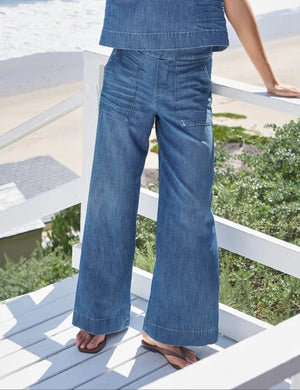 A person wearing FRANK & EILEEN’s Madeline Patch Pocket Pant in Famous Denim Distressed Wash stands on a white wooden deck overlooking greenery, sand, and the ocean, with only their lower body visible.