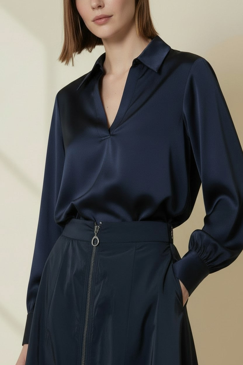 A person is wearing the VICARIO CINQUE - SOLEI TAFFETA TOP IN MIDNIGHT BLUE, a sleek V-neck blouse with long sleeves by VICARIO CINQUE, paired with a high-waisted skirt featuring a front zipper and O-ring pull. Reminiscent of the brand’s signature elegant style, the image captures the outfit from shoulders to hips.