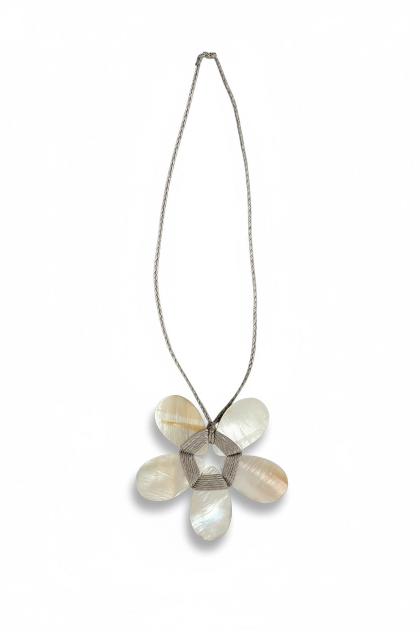A cream-colored mannequin showcases a textured beige jacket paired with the ANN LIGHTFOOT - FLAT MOTHER OF PEARL FLOWER NECKLACE, highlighting a striking white-petaled flower pendant with a metallic center suspended from an adjustable braided cord. The background is softly blurred.
