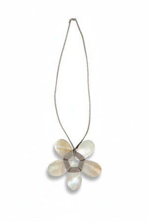 A cream-colored mannequin showcases a textured beige jacket paired with the ANN LIGHTFOOT - FLAT MOTHER OF PEARL FLOWER NECKLACE, highlighting a striking white-petaled flower pendant with a metallic center suspended from an adjustable braided cord. The background is softly blurred.