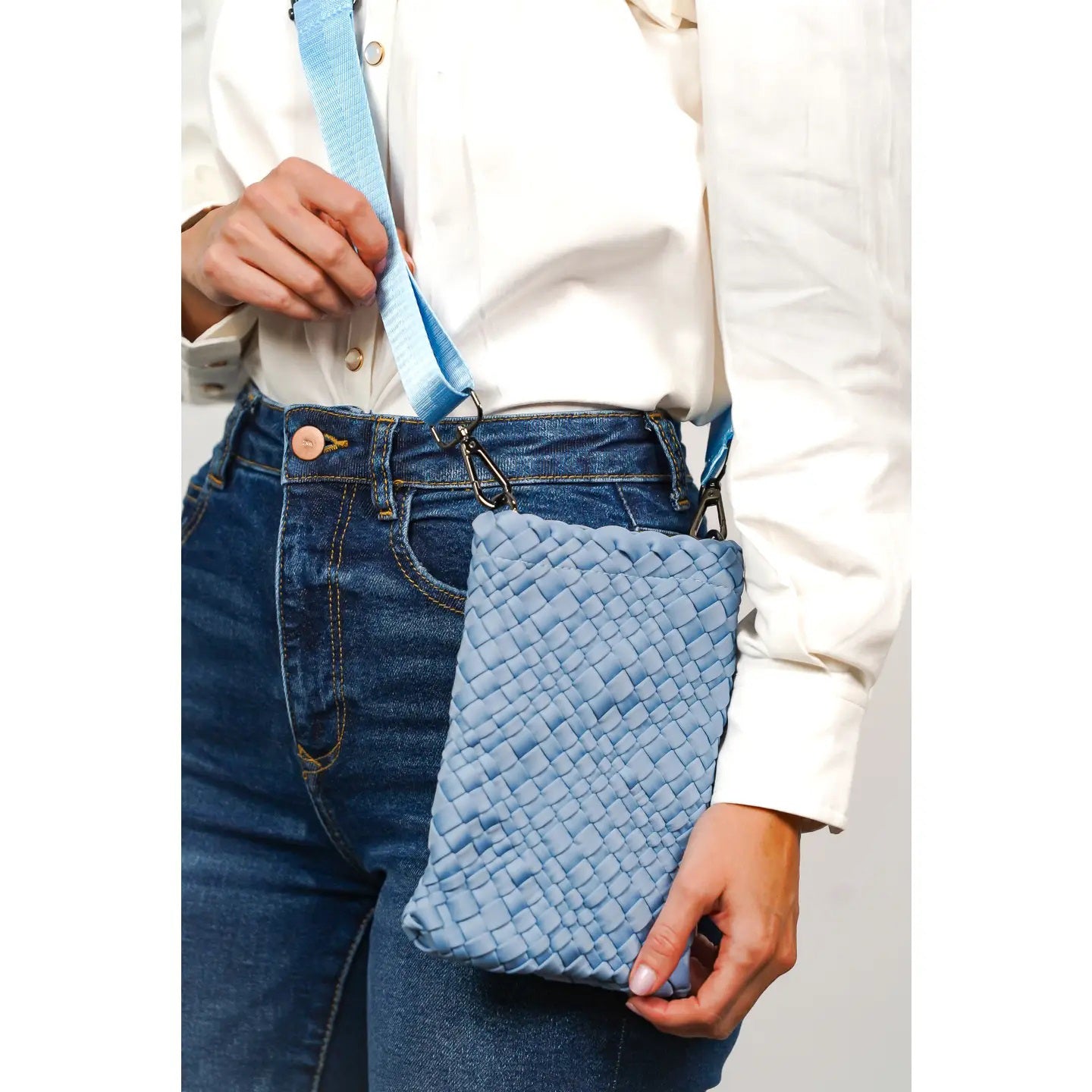 A person wearing blue jeans and a white shirt holds a light blue woven crossbody bag with a matching blue strap. The bag hangs at their hip.