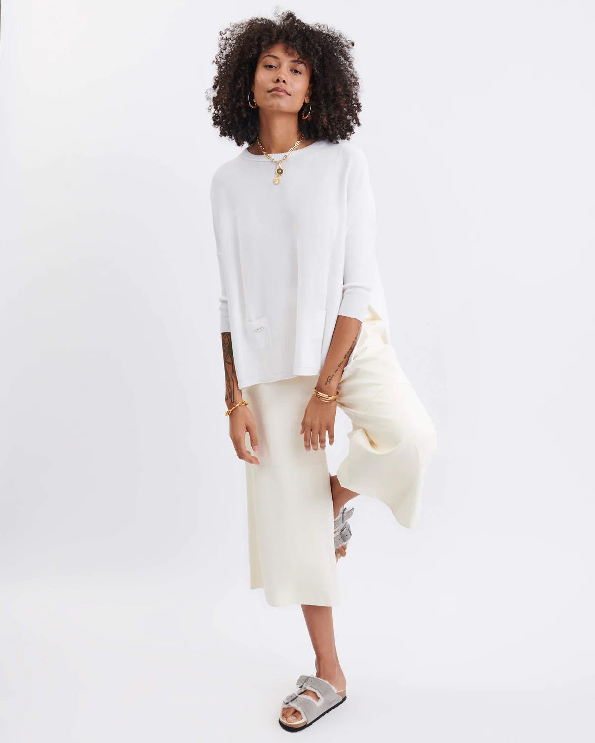 A smiling woman with curly hair wears the MERSEA - CATALINA CREWNECK SWEATER in navy blue and jeans, standing against a plain light background with her hands near her head.