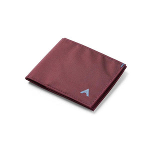 An ALLETT Sport Wallet in RFID-blocking mulberry nylon, along with a pen, sunglasses, candle, seashell, iPhone, and AirPods are neatly displayed on a marble surface.