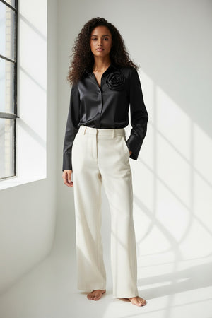 A woman with dark hair and a neutral expression wears the HINSON WU HALSEY Long Sleeve Cotton Satin Shirt with a large flower detail on the shoulder and black trousers, standing against a plain background.