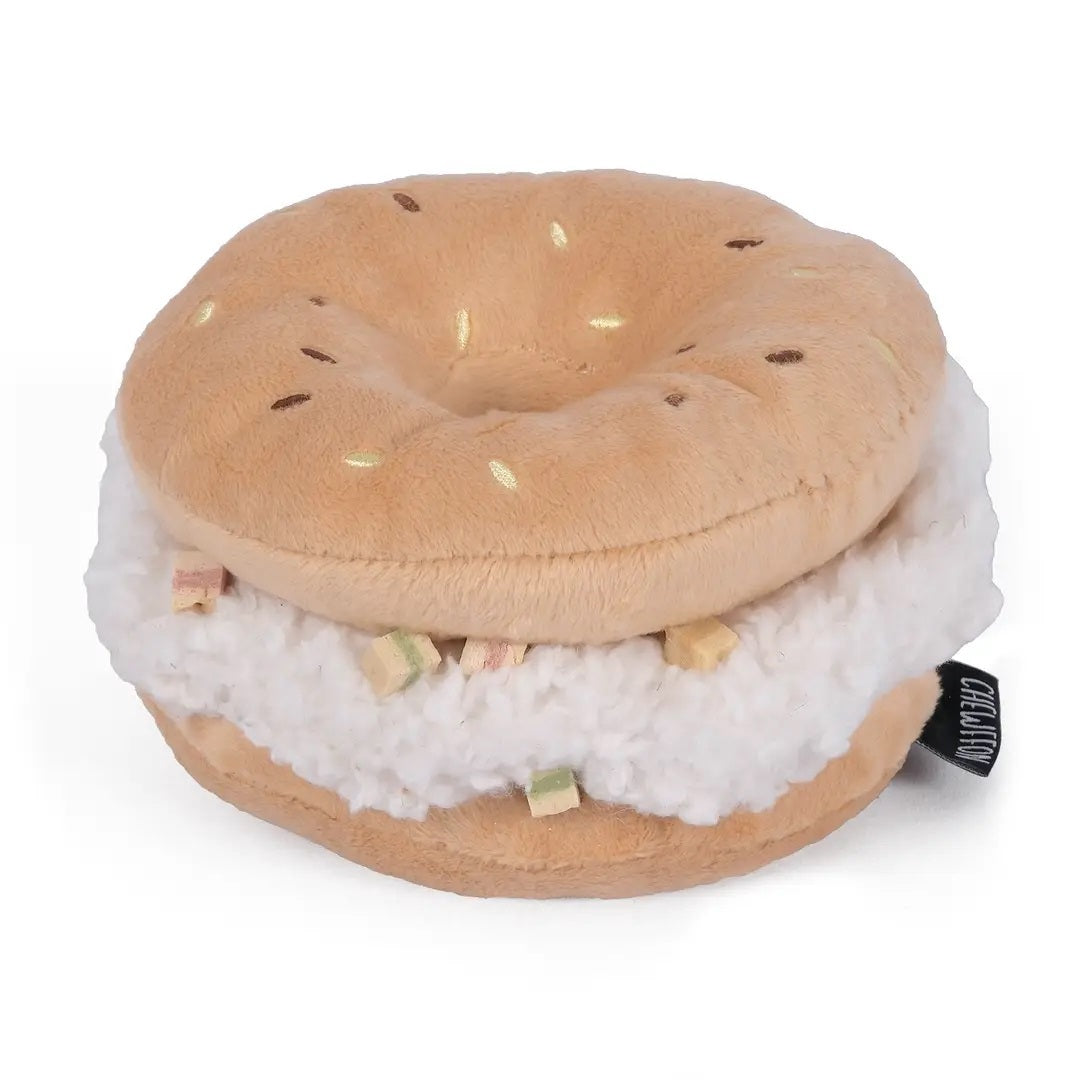 A small, fluffy white dog sniffs the CHEWFFON BAGEL PLUSH SQUEAKY DOG TOY, a large, round, beige plush toy resembling a sandwich, set against a plain light beige background.