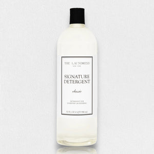 A 32 fl oz (946 ml) bottle of THE LAUNDRESS - SIGNATURE DETERGENT 32OZ featuring a classic scent and bio-based ingredients. The clear bottle comes with a black screw-top lid and a white label showcasing the product name and brand in black text. The background is a simple light gray.