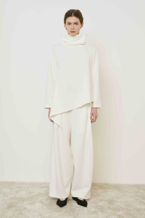 A person with brown hair faces away wearing the MEIMEIJ ASYMMETRICAL COWLNECK TOP in lightweight scuba fabric, featuring an asymmetrical hem, paired with matching white bottoms, against a plain white background.