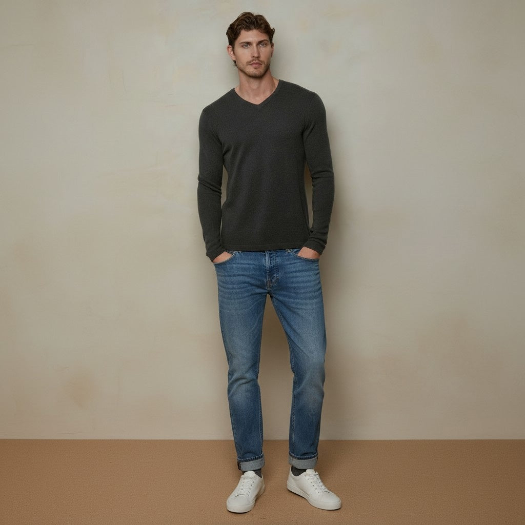 A man stands against a beige wall with his hands in his pockets, wearing the AUTUMN CASHMERE - Cashmere Basic V Neck with Open Bottom in Pepper by AUTUMN CASHMERE, paired with blue jeans and white sneakers.
