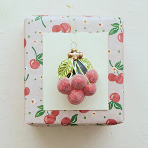 A GLASS CHERRY CLUSTER ORNAMENT WITH GLITTER and a green leaf hangs over a gift wrapped in cherry-patterned paper.