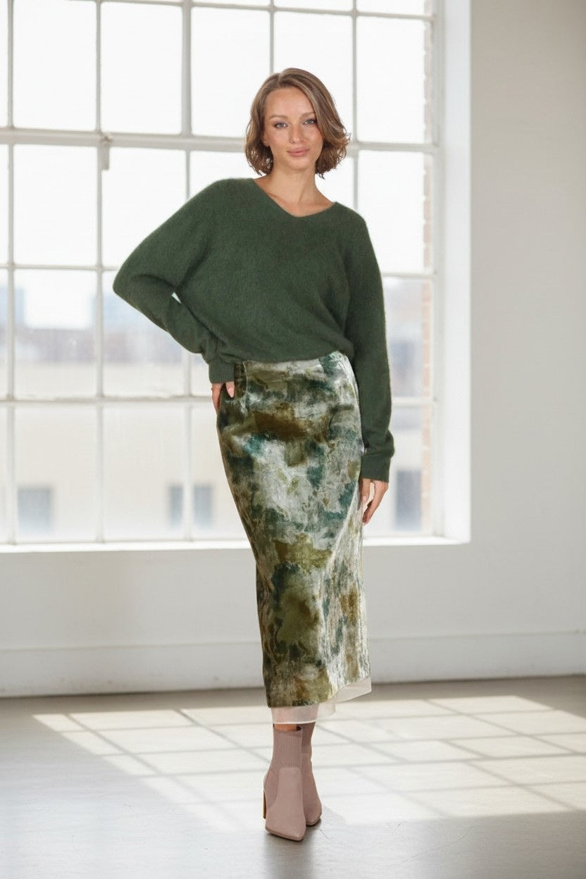 A woman stands indoors by a large window, smiling with one hand on her hip, wearing a fuzzy green sweater, light-colored ankle boots, and the JESSIE LIU Printed Velvet Midi Skirt with Organza Trim by JESSIE LIU—a green and beige tie-dye skirt featuring a long straight silhouette.