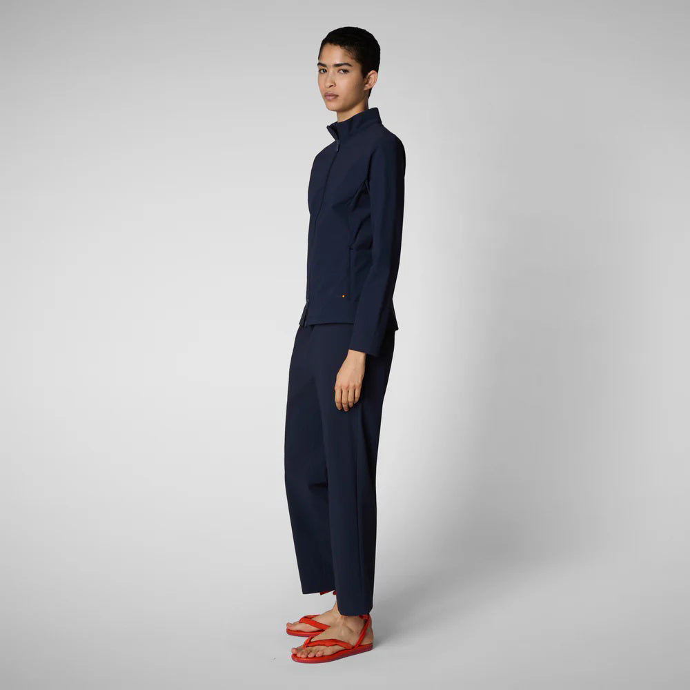 A person with shoulder-length hair and a neutral expression wears a white long-sleeve shirt, SAVE THE DUCK MILAN PULL ON PANT in black wrinkle-free fabric, and black slip-on shoes. Their hands rest at their sides against a plain background.