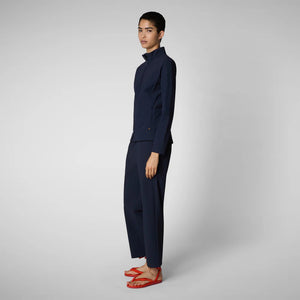 A person with shoulder-length hair and a neutral expression wears a white long-sleeve shirt, SAVE THE DUCK MILAN PULL ON PANT in black wrinkle-free fabric, and black slip-on shoes. Their hands rest at their sides against a plain background.