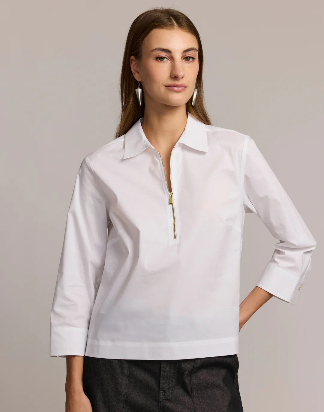 A woman with long brown hair wears the HINSON WU - ALESSA 3/4 SLEEVE SHIRT in olive green, featuring a zip-up collar and stretch polished cotton blend, paired with dark pants. She stands against a light background with minimal makeup and geometric earrings.