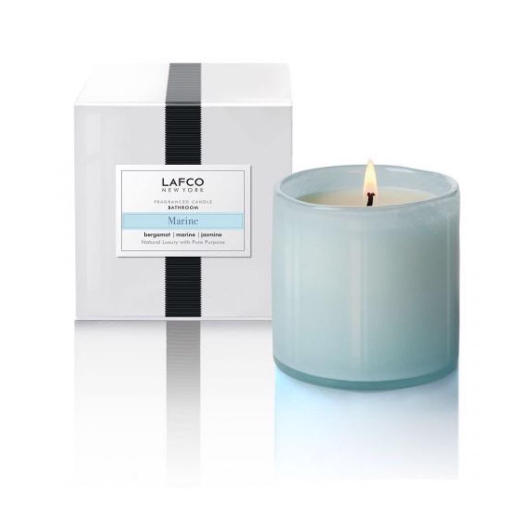A LAFCO - MARINE, BATHROOM 150Z candle from LAFCO illuminates next to its packaging. The elegant candle is encased in a light blue glass holder, while the box is white with a black stripe and labeled with &quot;Marine, Bathroom, Bergamot, Marine, Jasmine.&quot; Hand-poured craftsmanship accentuates the calming water lily aroma.