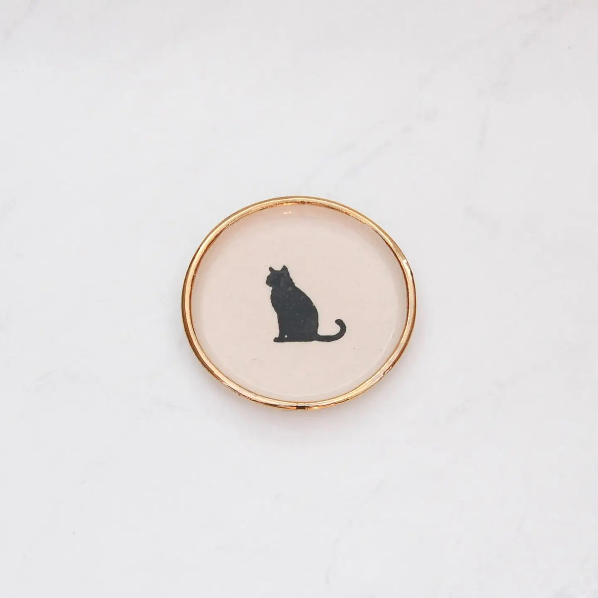 The COMPOST DESIGN Sitting Cat Tiny Dish 3&quot; features a black silhouette of a sitting cat on a cream background, framed by an elegant 22k gold edge. Perfect for decorative use, it rests beautifully on light, textured surfaces.