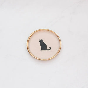 The COMPOST DESIGN Sitting Cat Tiny Dish 3" features a black silhouette of a sitting cat on a cream background, framed by an elegant 22k gold edge. Perfect for decorative use, it rests beautifully on light, textured surfaces.