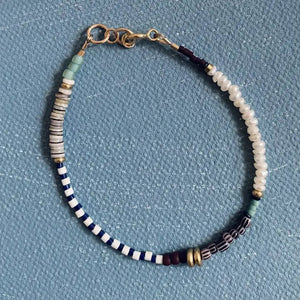 The ALICE RISE - PEARL BRACELET by ALICE RISE features delicate blue, white, green, cream, and gold beads in unique patterns with a 14k gold-filled clasp, shown on a textured blue surface.