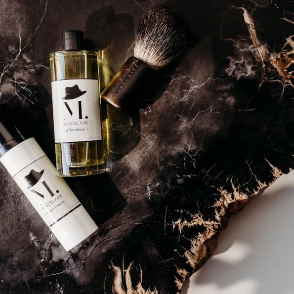 A clear bottle filled with yellow liquid, BAUDELAIRE - M.B. AFTERSHAVE by BAUDELAIRE features a classic design with a black hat graphic on the front and a black cap, offering a refreshing green-forest scent enriched with soothing witch hazel.