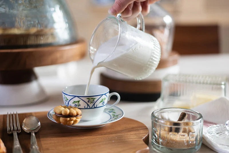 The ETUHOME - BISTRO GLASS MINI JUG by ETUHOME, filled with milk, rests on a white surface next to a butter dish and knife, with a clear glass baking dish and wooden container in the background.