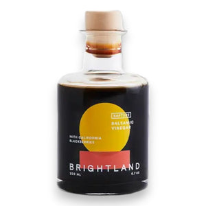 A 200 ml bottle of BRIGHTLAND - 'RAPTURE' Blackberry Balsamic Vinegar made with California Zinfandel grapes and triple crown blackberries. The bottle has a round body, a short neck, and is sealed with a light-colored cork stopper. Its label boasts a modern design featuring a yellow and orange circle against a black background.