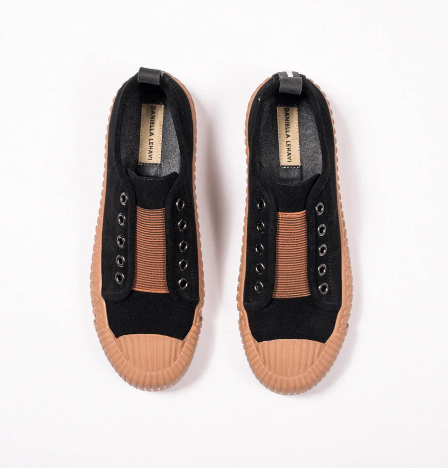 Introducing the DANIELLA LEHAVI - OSAKA SNEAKER by DANIELLA LEHAVI: Black sneakers featuring brown rubber soles and decorative brown straps across the top. They highlight a contemporary design with circular metal eyelets on the sides and are presented on a white platform against a simple, light-colored background.