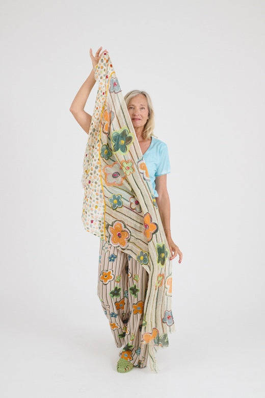 A woman with blonde hair stands against a white background, wearing the STORIATIPIC ANNE Scarf in beige featuring pastel stripes and floral prints with hand-embroidered linen details, paired with matching pants and green sandals.