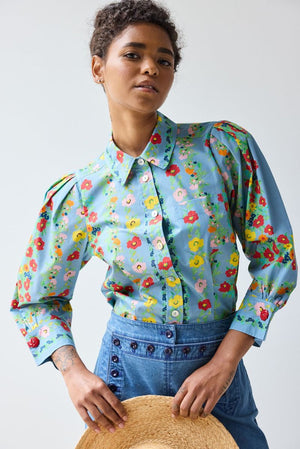 A young woman with curly hair looks over her shoulder, wearing the HOPE FOR FLOWERS - GIRL SHIRT IN CHAMBRAY FLORAL by HOPE FOR FLOWERS and high-waisted blue jeans with a lace-up back detail.