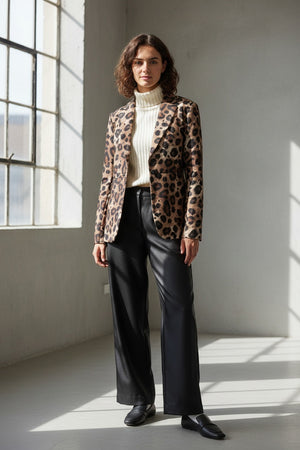 A person with wavy brown hair models the LEOPARD PRINT JACKET by ALYKI LABORATORIO over a white turtleneck sweater, standing with arms crossed against a plain white background.