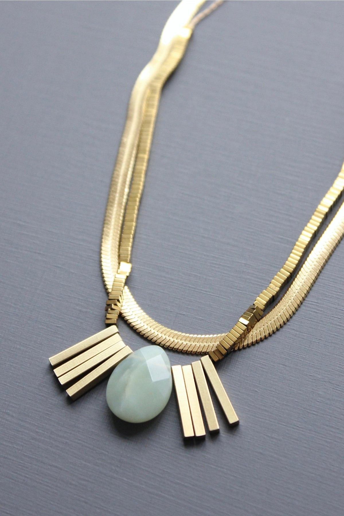 The DAVID AUBREY DOUBLE STRAND SNAKE CHAIN AND JADE ARTDECO PENDANT NECKLACE features two gold-tone layered chains, a large oval jade pendant, and four small gold rectangular accents, beautifully displayed on a gray background.
