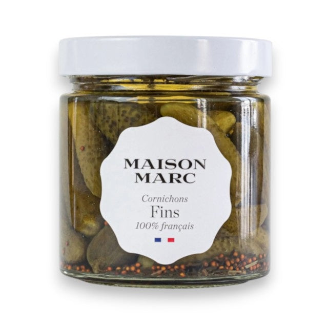 A glass jar of FRENCH FEAST MAISON MARK - EXTRA FINE CORNICHONS FINS, featuring a white label that reads “Cornichons Fins 100% français,” has a white lid and contains small Burgundy gourmet pickles in brine.