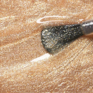 A hand with medium-brown skin displays short, neatly manicured nails painted with MANUCURIST - GREEN™ NAIL POLISH IN GOLD by MANUCURIST INC, featuring a shimmery pale gold glitter finish against a neutral beige background.