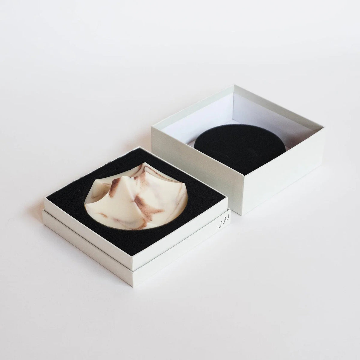 The UME STUDIO UME - LAVENDER CREME ERODE SOAP, with white and reddish-brown swirls and sculpted peaks, rests on a round marble-patterned soap dish against a plain background.