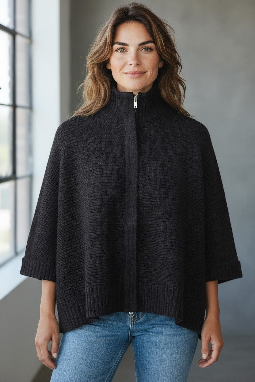 A woman with wavy brown hair stands indoors by a sunlit window, wearing the KINROSS CASHMERE Rib Zip Mock Sleeveless Cashmere Cardigan in black paired with light blue jeans.