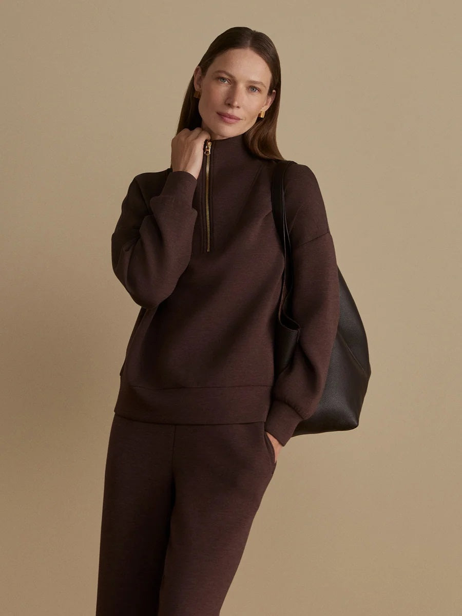 A person wears an oversized, dark brown VARLEY - HAWLEY HALF-ZIP SWEATSHIRT IN COFFEE BEAN MARL with a gold zipper.