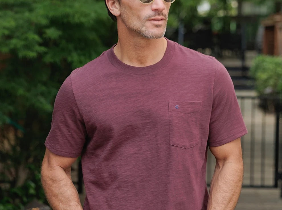 A person wearing the CRIQUET Slub Cotton Short Sleeve Pocket T-Shirt in Jam Band, featuring a small chest pocket and a subtle blue logo. The blurred greenery and wood background perfectly complement this vintage-inspired CRIQUET shirt, creating an effortlessly classic look.