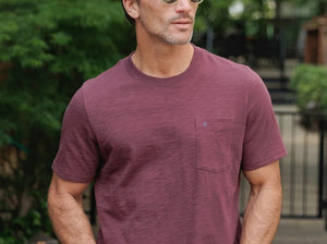 A person wearing the CRIQUET Slub Cotton Short Sleeve Pocket T-Shirt in Jam Band, featuring a small chest pocket and a subtle blue logo. The blurred greenery and wood background perfectly complement this vintage-inspired CRIQUET shirt, creating an effortlessly classic look.
