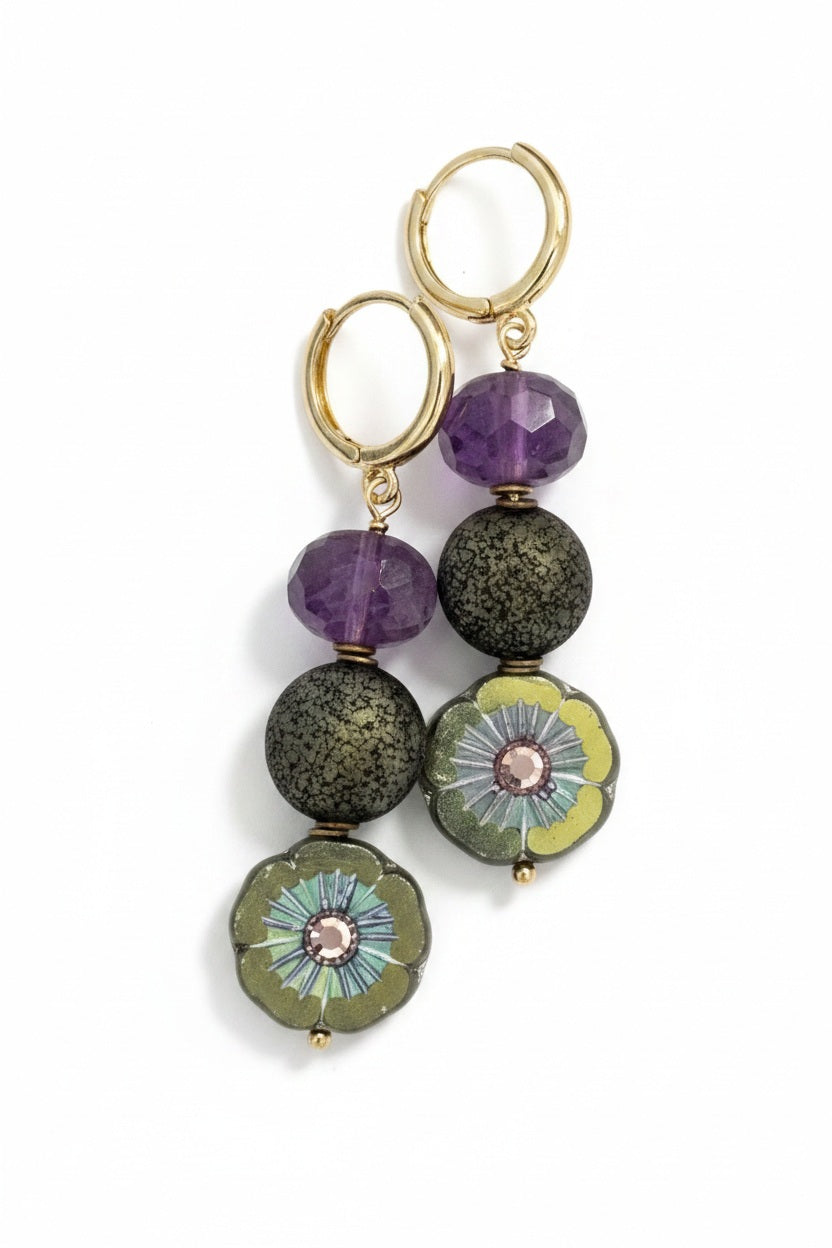 The SANDRINE FRANCE STUDIO - BEIRA EARRINGS by SANDRINE FRANCE STUDIO showcase gold hoops adorned with purple faceted beads, textured round beads, and flat floral beads in green and blue with a pink center, all elegantly arranged in a vertical design against a white background.