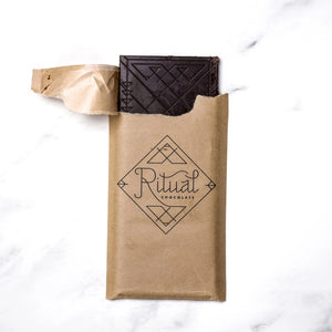 A gold and yellow RITUAL CHOCOLATE - HONEYCOMB TOFFEE CHOCOLATE BAR wrapper with geometric designs holds a 2.12 oz (60 g), 75% cacao single origin dark chocolate bar, displayed on a marble background.