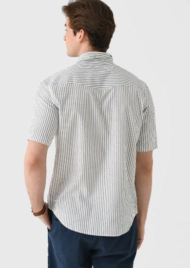 A young man, smiling with one hand in his pocket, wears CRIQUET’s Short Sleeve Performance Seersucker Pearl Snap Shirt in Sage and navy blue pants.