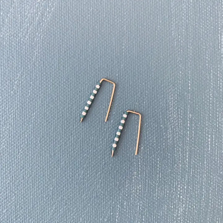 The ALICE RISE - LITTLE QUILL EARRINGS IN SLATE BLUE STRIPE feature unique 14k gold filled posts adorned with alternating white and green beads, arranged parallel on a textured light gray surface.
