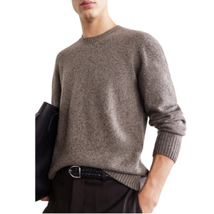 A person wearing the HARTFORD Seamless Wool Crew Pullover in Mushroom, an Italian-made sweater, paired with dark pants, is seated with one arm resting on their knee against a plain light background.