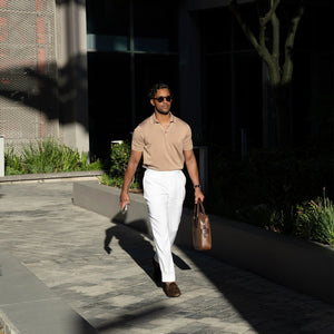 A man in sunglasses wears the COLLARS AND CO MARANELLO Mid-Weight Cotton & Silk 3-Button Polo in Taupe with white trousers, carrying a brown leather bag and phone. Green plants and modern buildings are in the background.