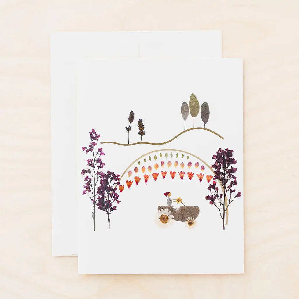 The PETAL PEOPLE PRESS - FARMER IN THE DELL CARD by PETAL PEOPLE PRESS features a car driving under a rainbow of hearts, surrounded by flowers, trees, and hills—an ideal all-occasion card for any nature lover.