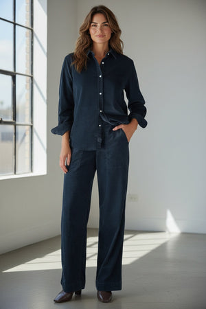 A person stands indoors by a window, dressed in the FRANK & EILEEN MADELEINE Patch Pocket Pull On Pant in Italian Corduroy Classic Navy and a coordinating dark navy button-up shirt, paired with brown shoes. With one hand tucked into their pocket, they smile softly as sunlight streams into the minimalist room.