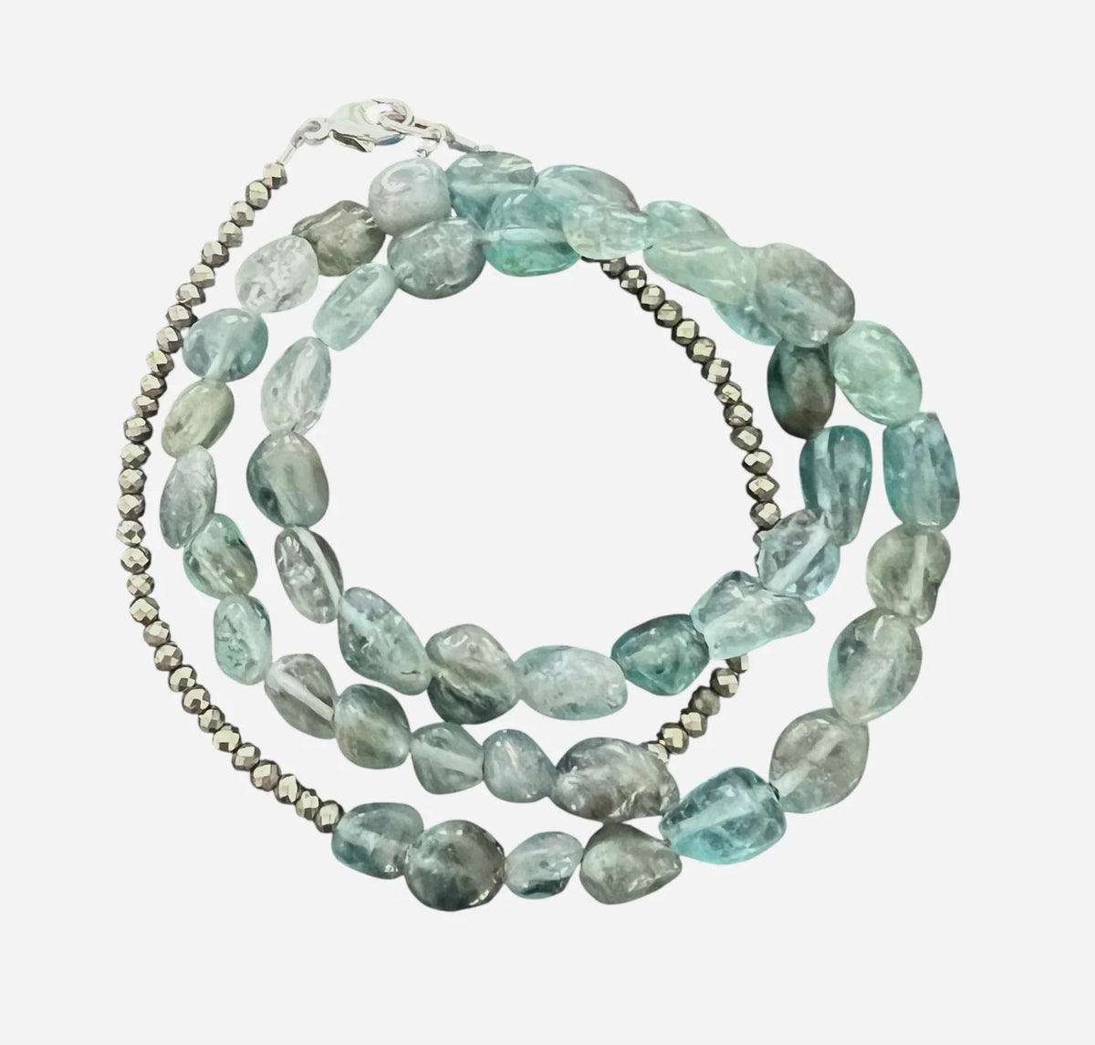 The ANN LIGHTFOOT - APATITE NUGGET TRIPLE WRAP BRACELET by ANN LIGHTFOOT features irregular light blue-green apatite stones, metallic round beads, and a sterling silver clasp, coiled in a circle on a dark surface.