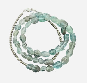 The ANN LIGHTFOOT - APATITE NUGGET TRIPLE WRAP BRACELET by ANN LIGHTFOOT features irregular light blue-green apatite stones, metallic round beads, and a sterling silver clasp, coiled in a circle on a dark surface.